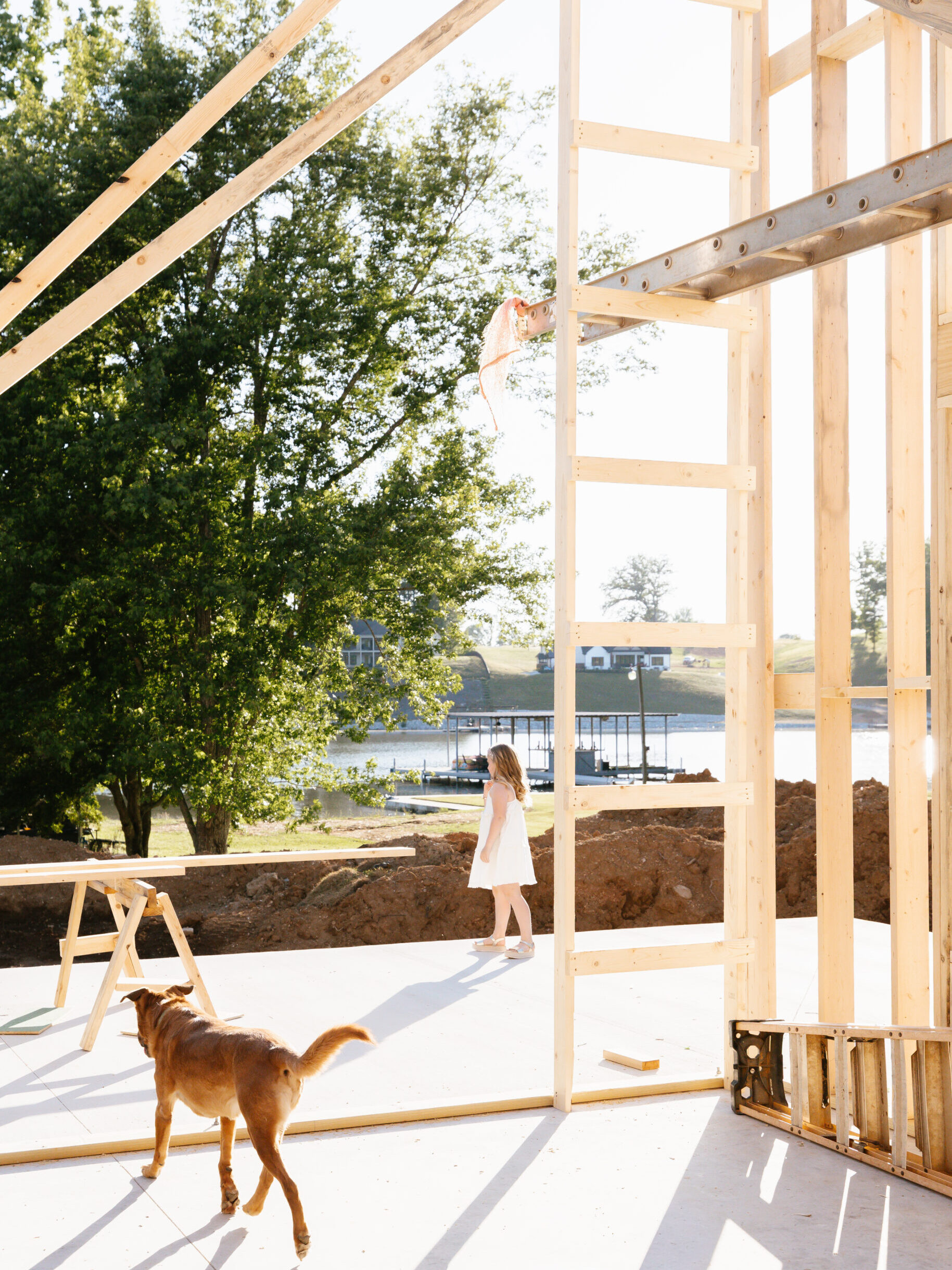 Girl in a white dress stands beside a wooden-framed building under construction; a brown dog walks nearby.