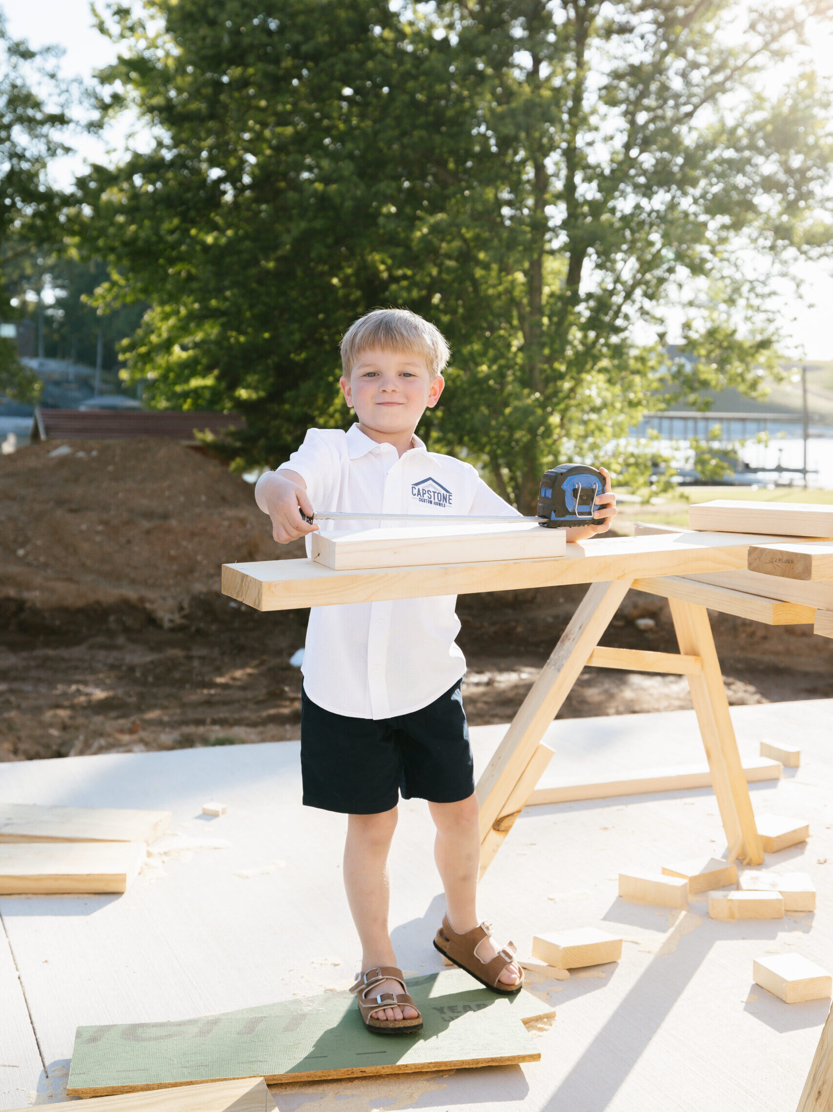 Young boy in a white shirt and black shorts holds a tape measure at a wooden workbench on a construction site outdoors.