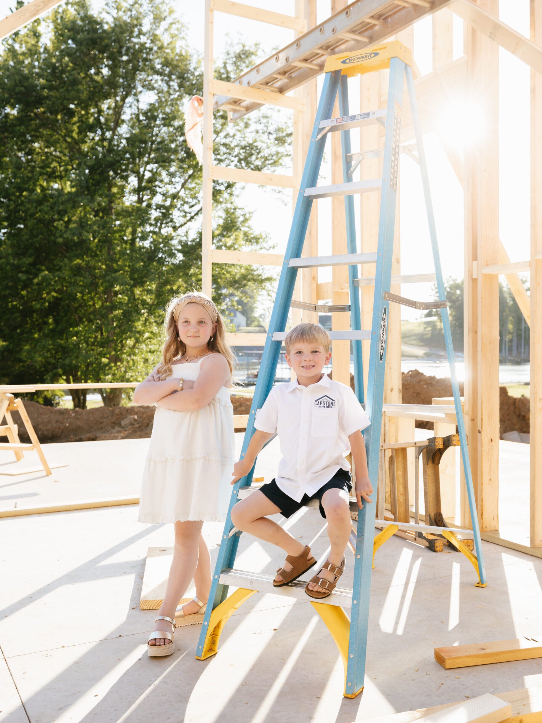 Two children stand at a construction site with a blue ladder and wooden framing behind them.