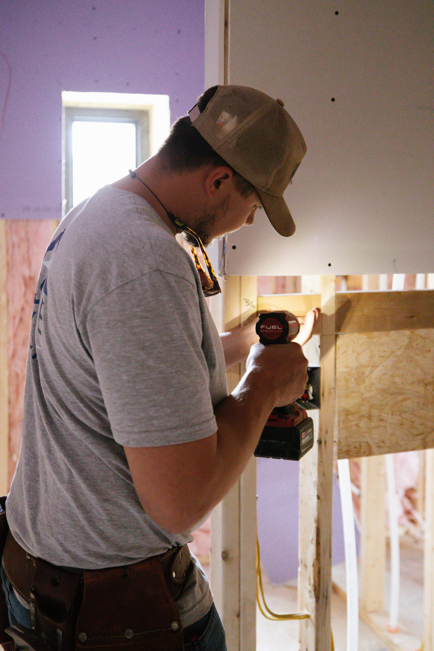 Man in a cap and gray shirt drills into wooden studs in a partially framed room, safety glasses hanging from his neck.