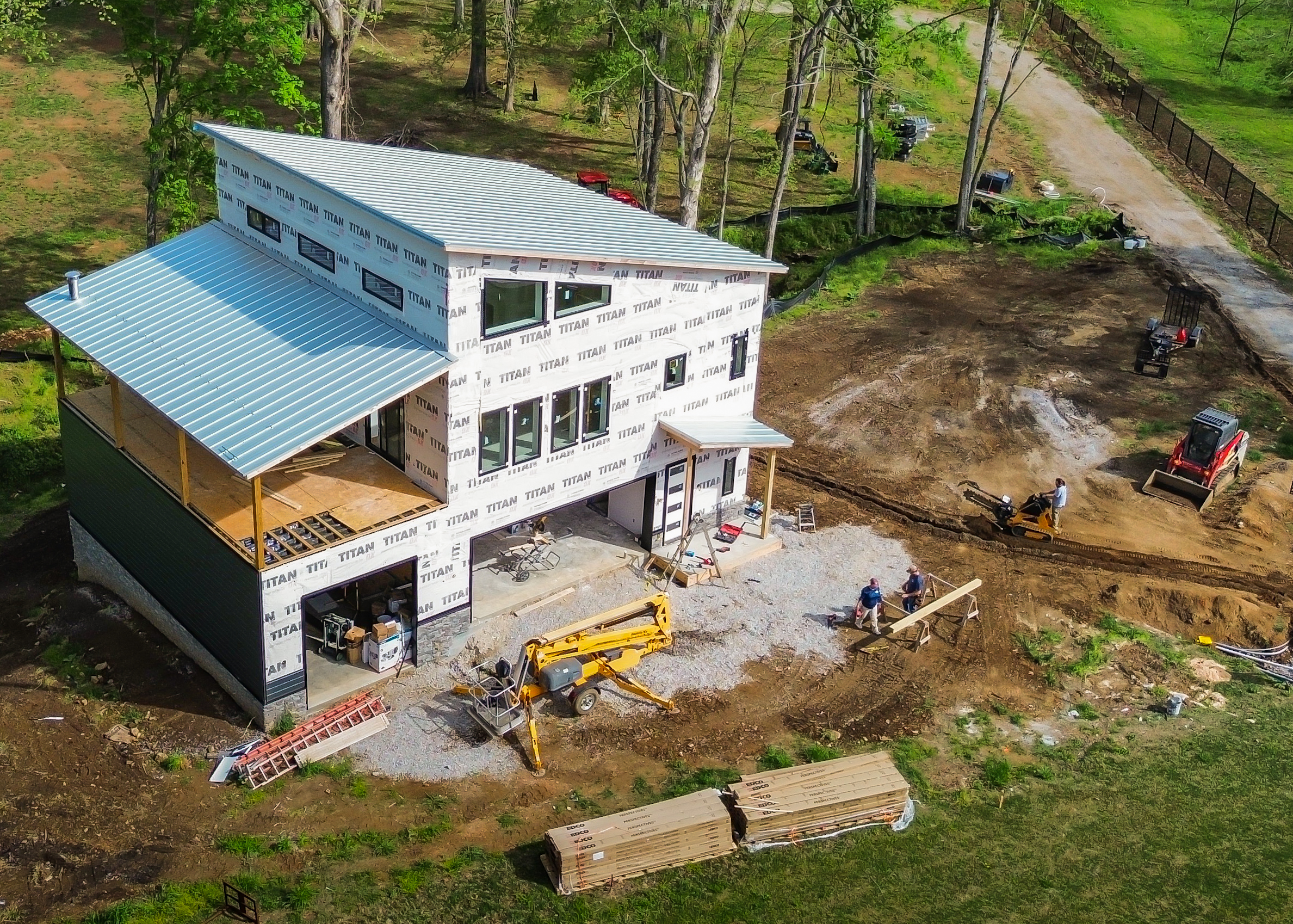 A partially built two-story home with a silver metal roof and Titan-wrapped walls in a wooded construction site.