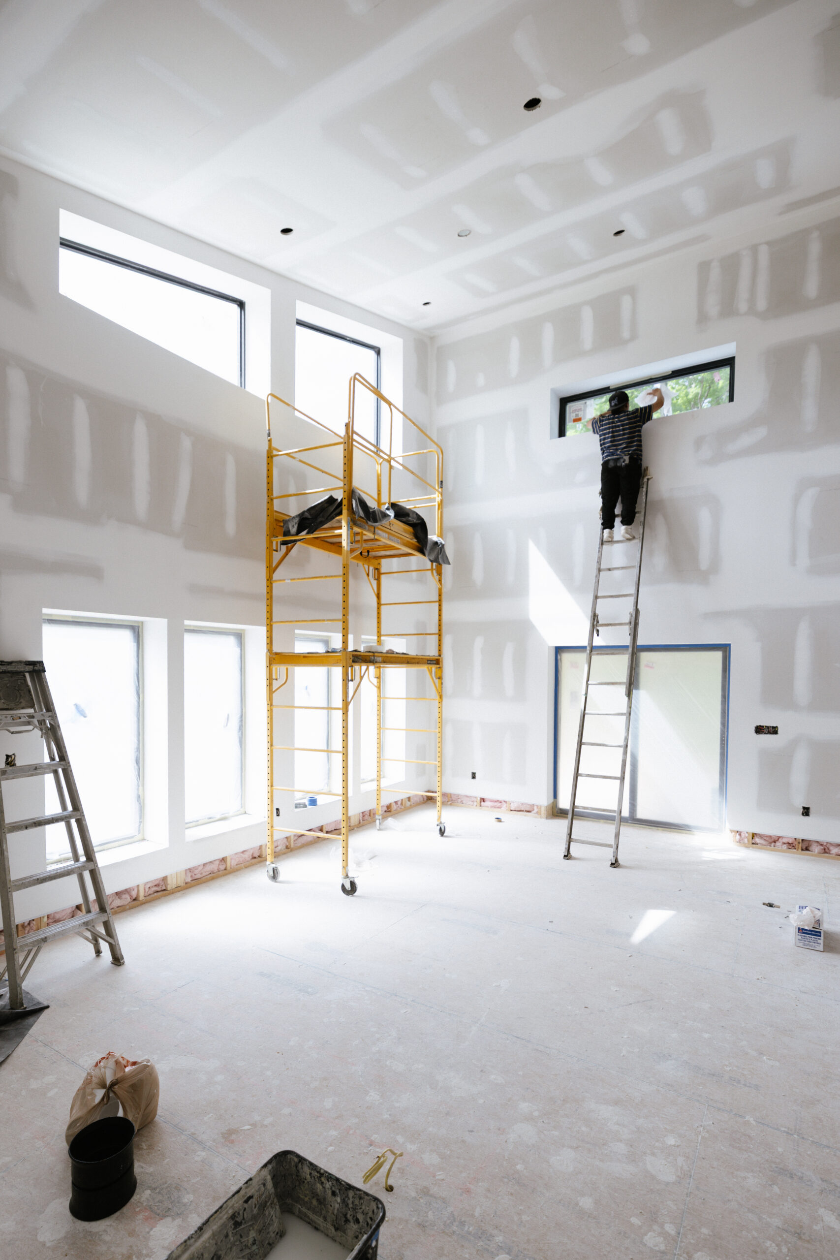 Interior of a building under construction with drywall seams, scaffolding, and a worker on a ladder near a window site clean-up in progress.