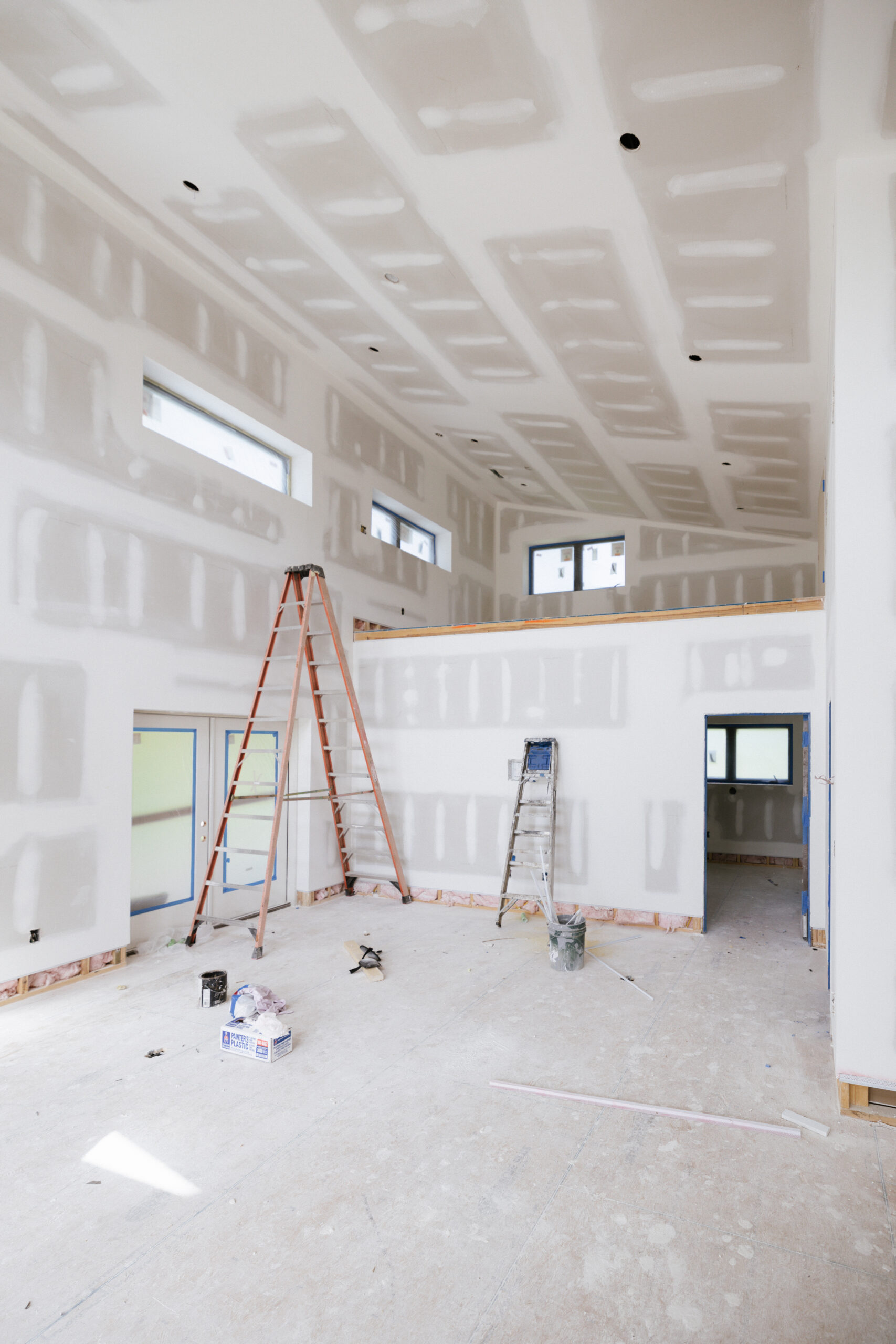 Interior of a large unfinished room with drywall installed and seams visible, ladders leaning against walls, and construction tools on the floor