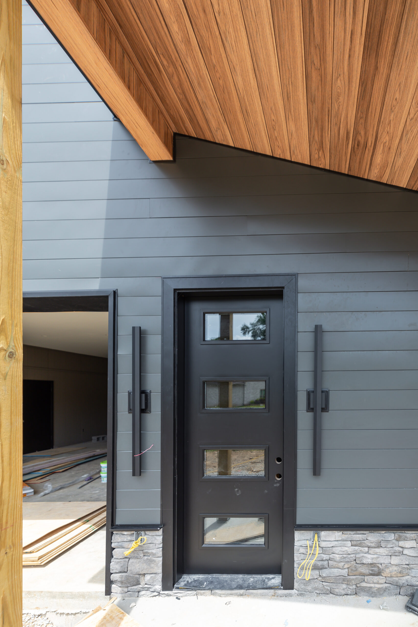 Modern entry door with four rectangular glass panes, dark frame, and light gray siding under a wooden overhang.