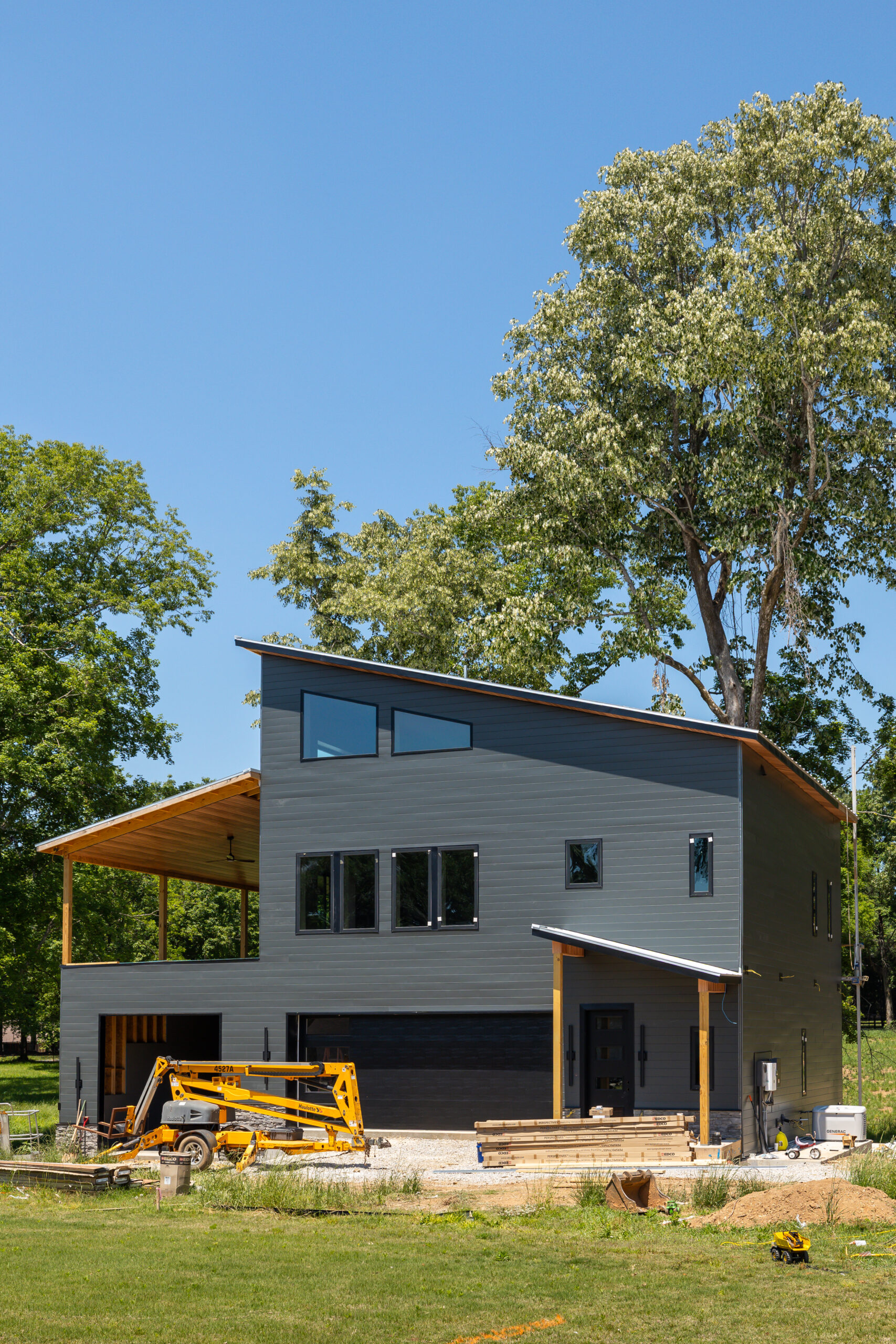 Modern two-story house under construction with dark gray siding, large windows, and wooden framing visible on the left; construction materials and equipment in the yard.