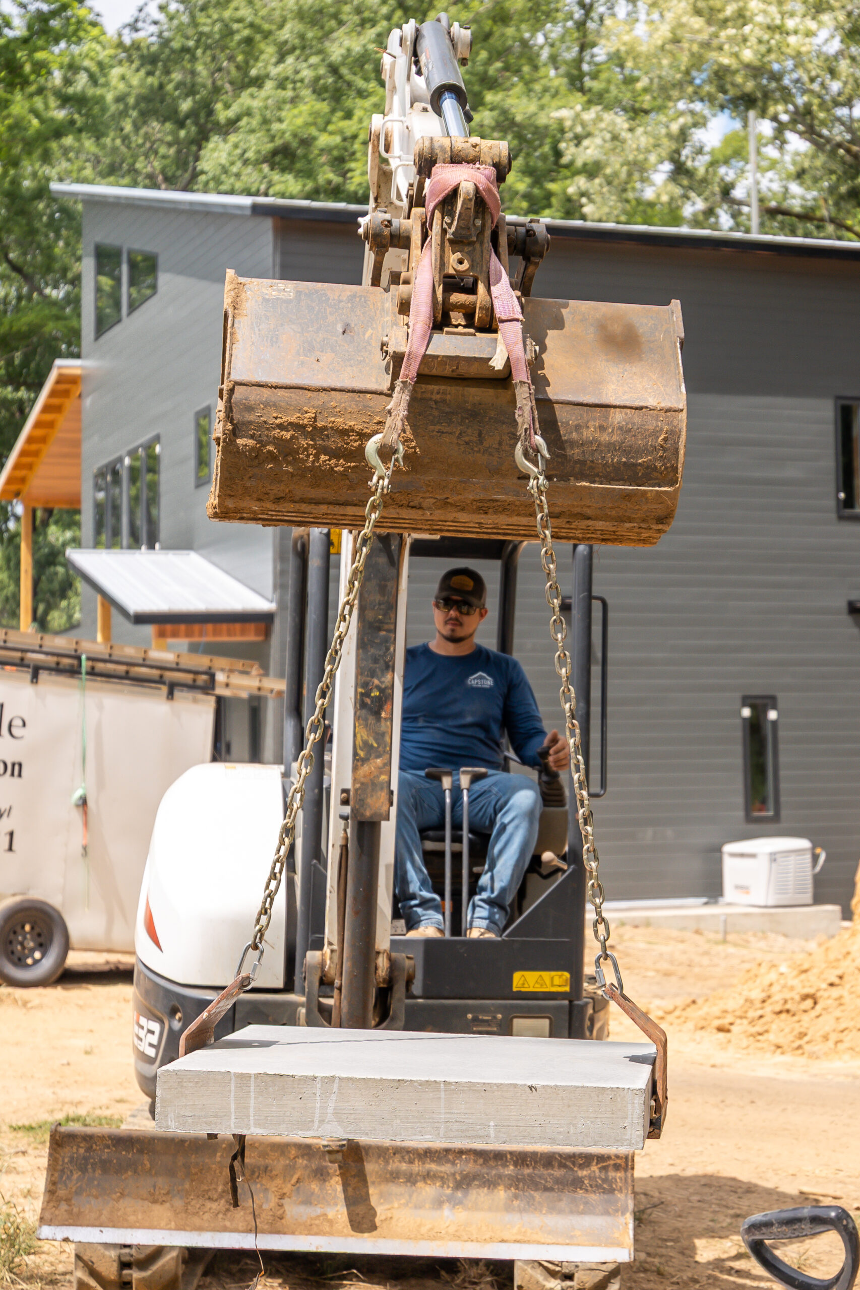 Operator seated in excavator cab controlling a large dirt-filled bucket raised on a construction site, with a gray building in the background