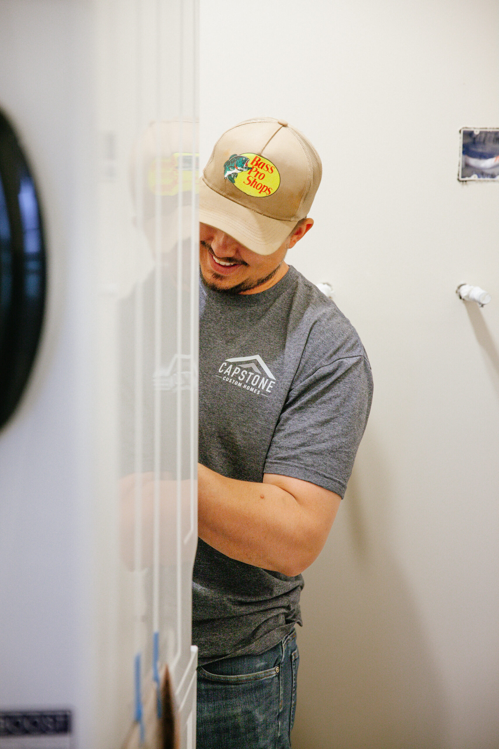 Man wearing a beige Bass Pro Shops cap and gray t‑shirt stands indoors, looking down beside a white cabinet/partition.