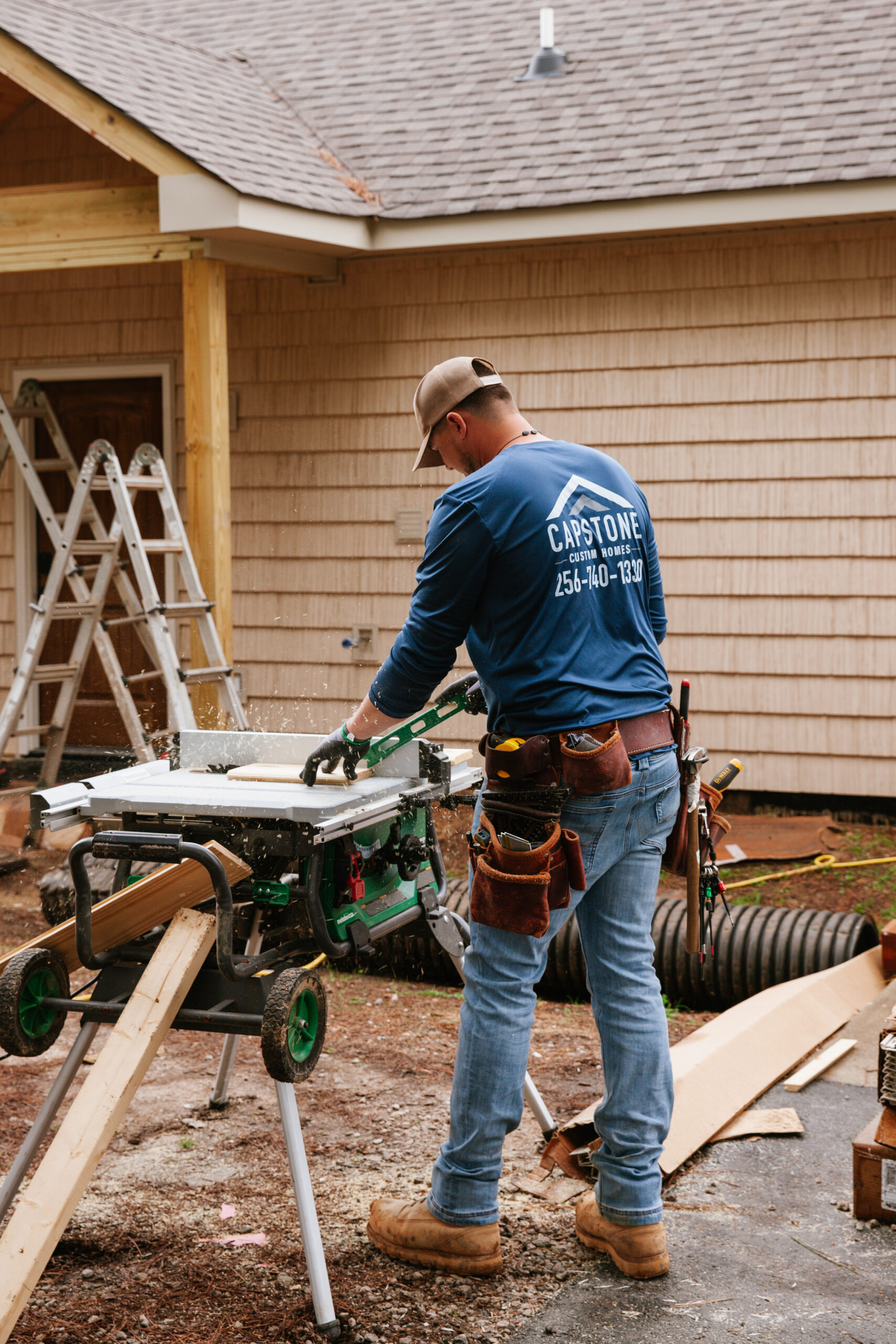 Carpenter wearing a cap and tool belt cuts wood on a table saw at an outdoor job site in front of a beige house outside renovation.