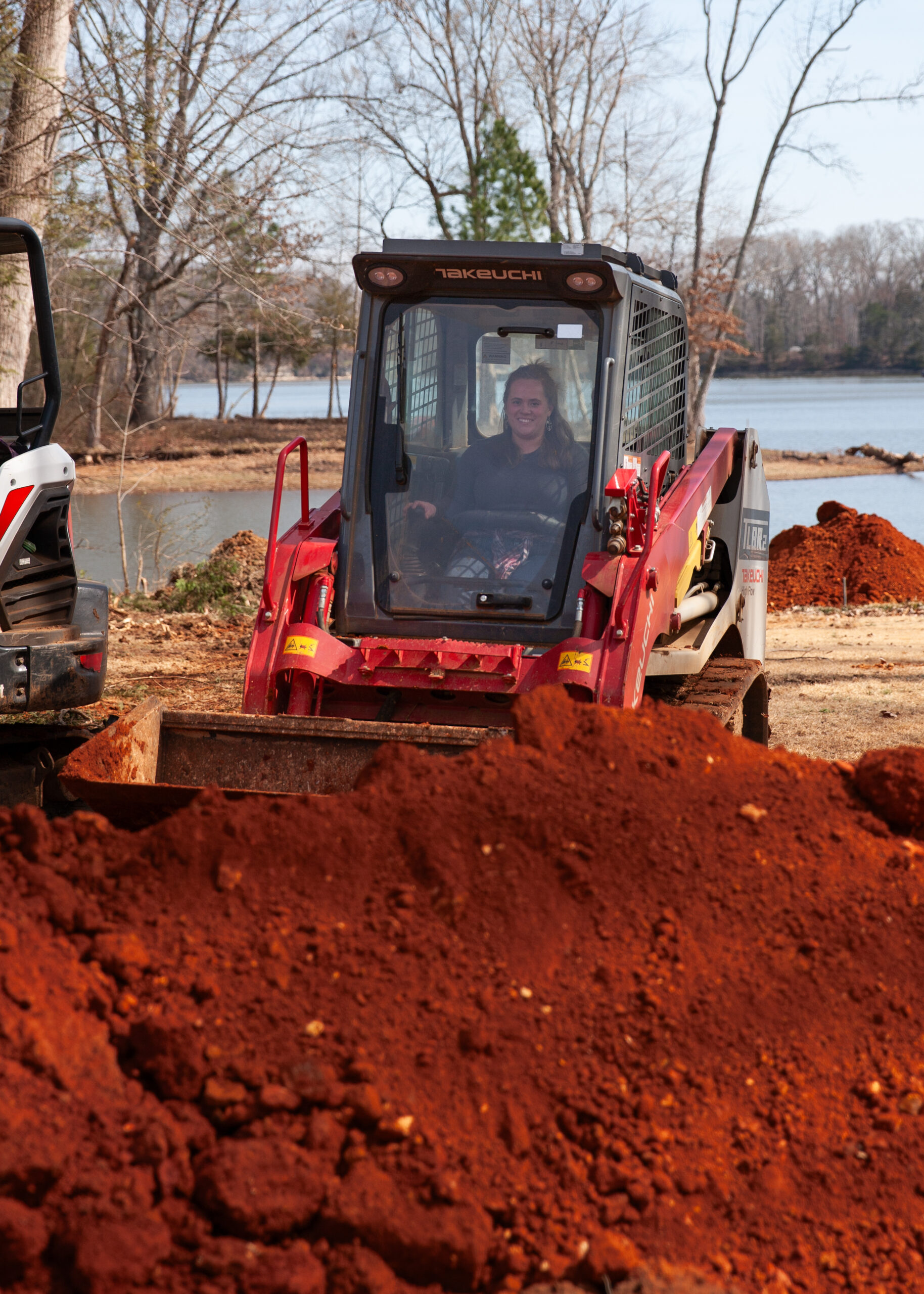 Woman sits in the cab of a red Takeuchi excavator on a construction site, with a mound of red earth and a lake in the background.