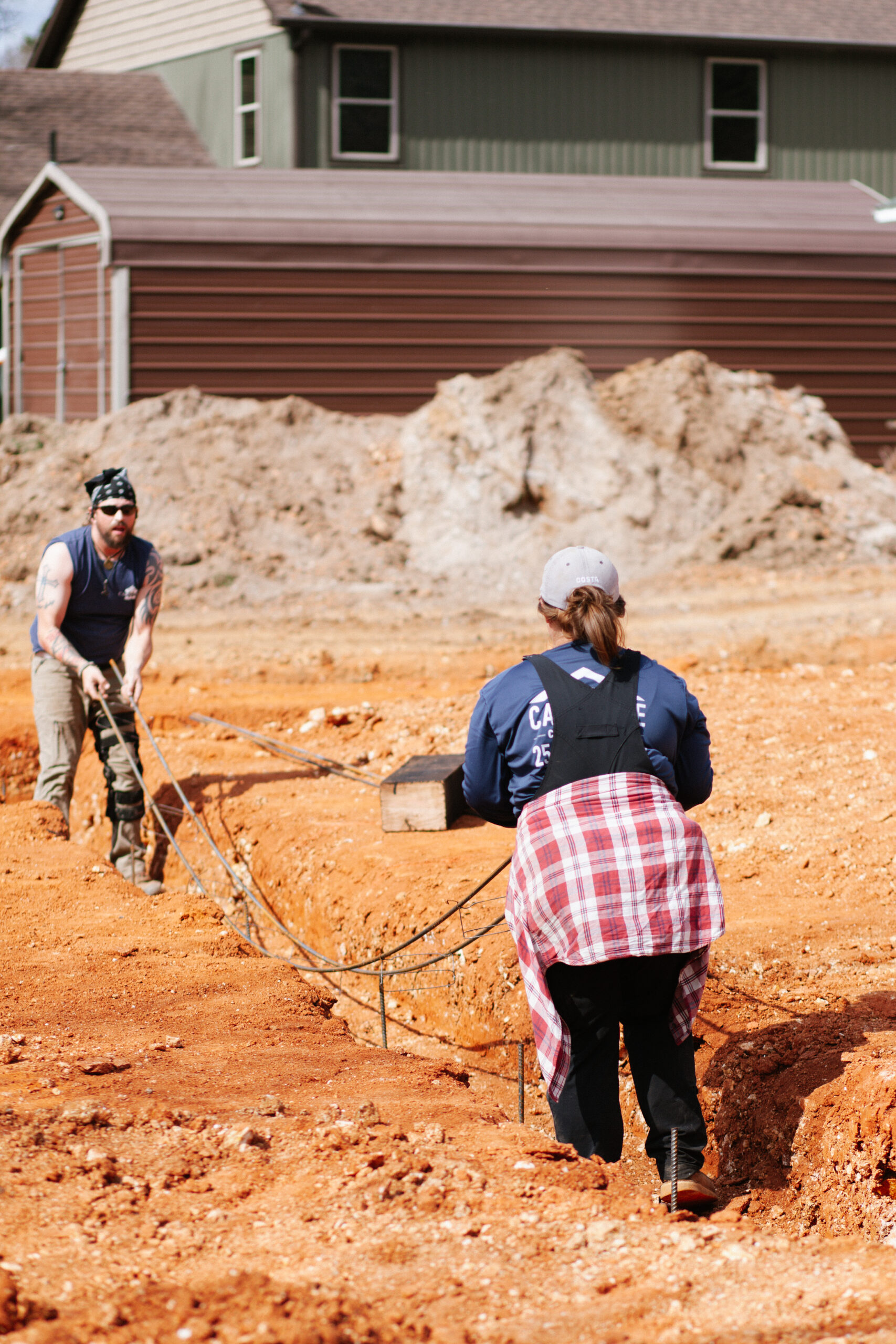 Two construction workers in a dirt excavation site with a large dirt pile and a house behind them; man with a bandana and sleeveless shirt operates a tool while a woman in a cap and plaid shirt assists.