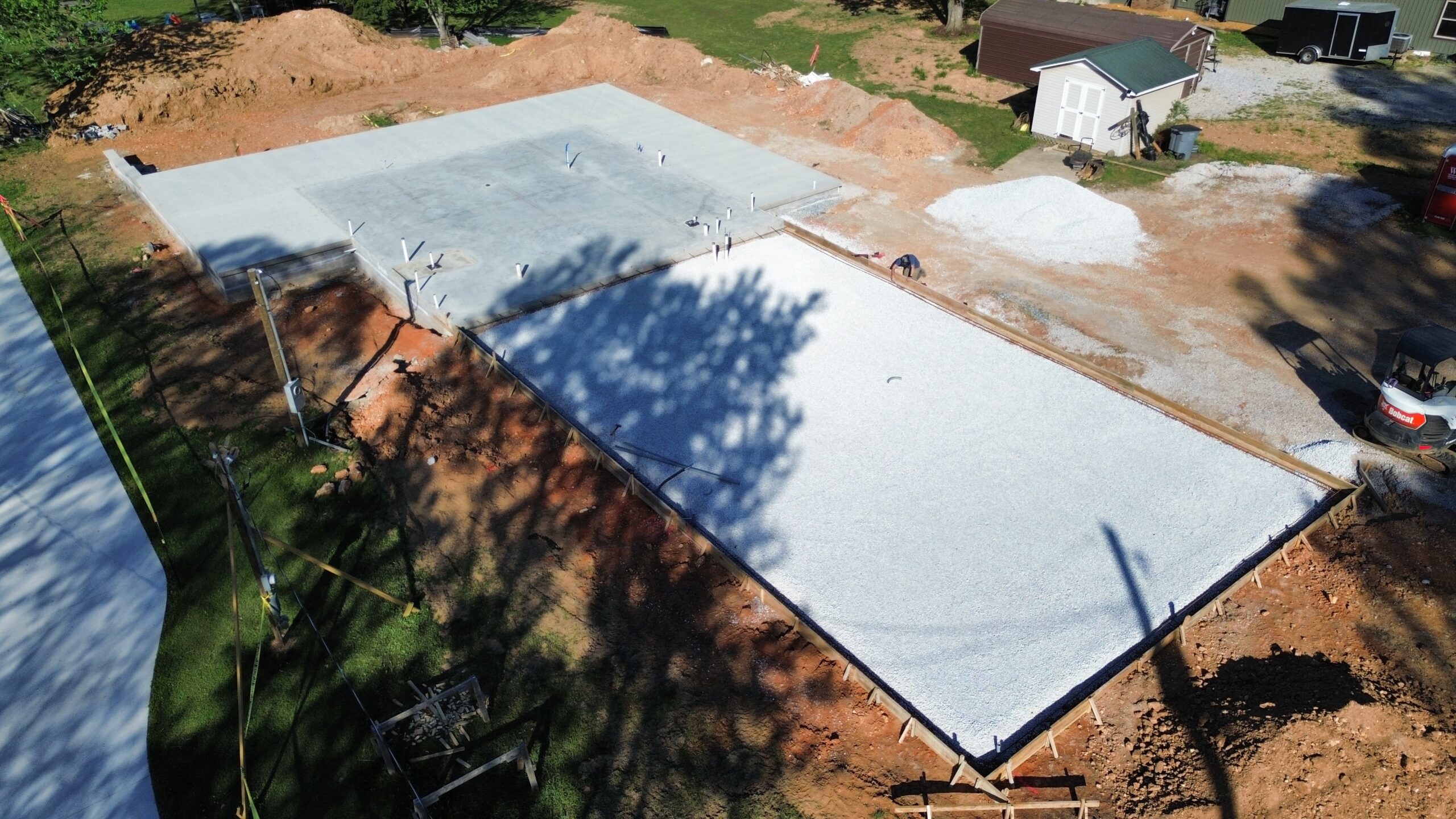Aerial view of a residential construction site with a concrete slab foundation and surrounding dirt piles and equipment nearby.