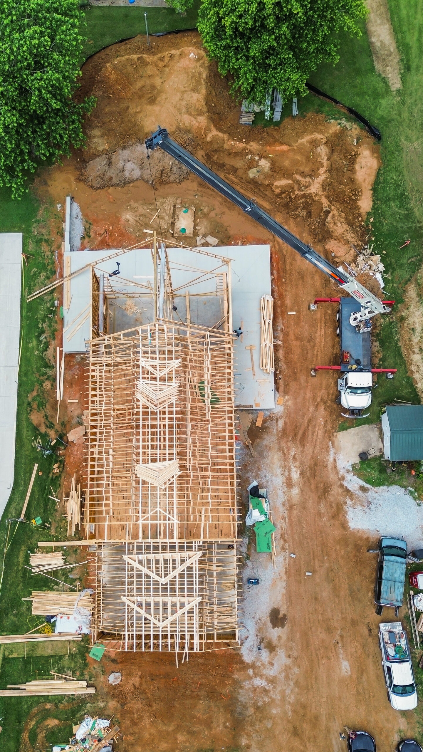 Aerial view of a construction site showing a wooden-framed building under construction, a large crane truck extending over the dirt yard, and piles of lumber around the structure.