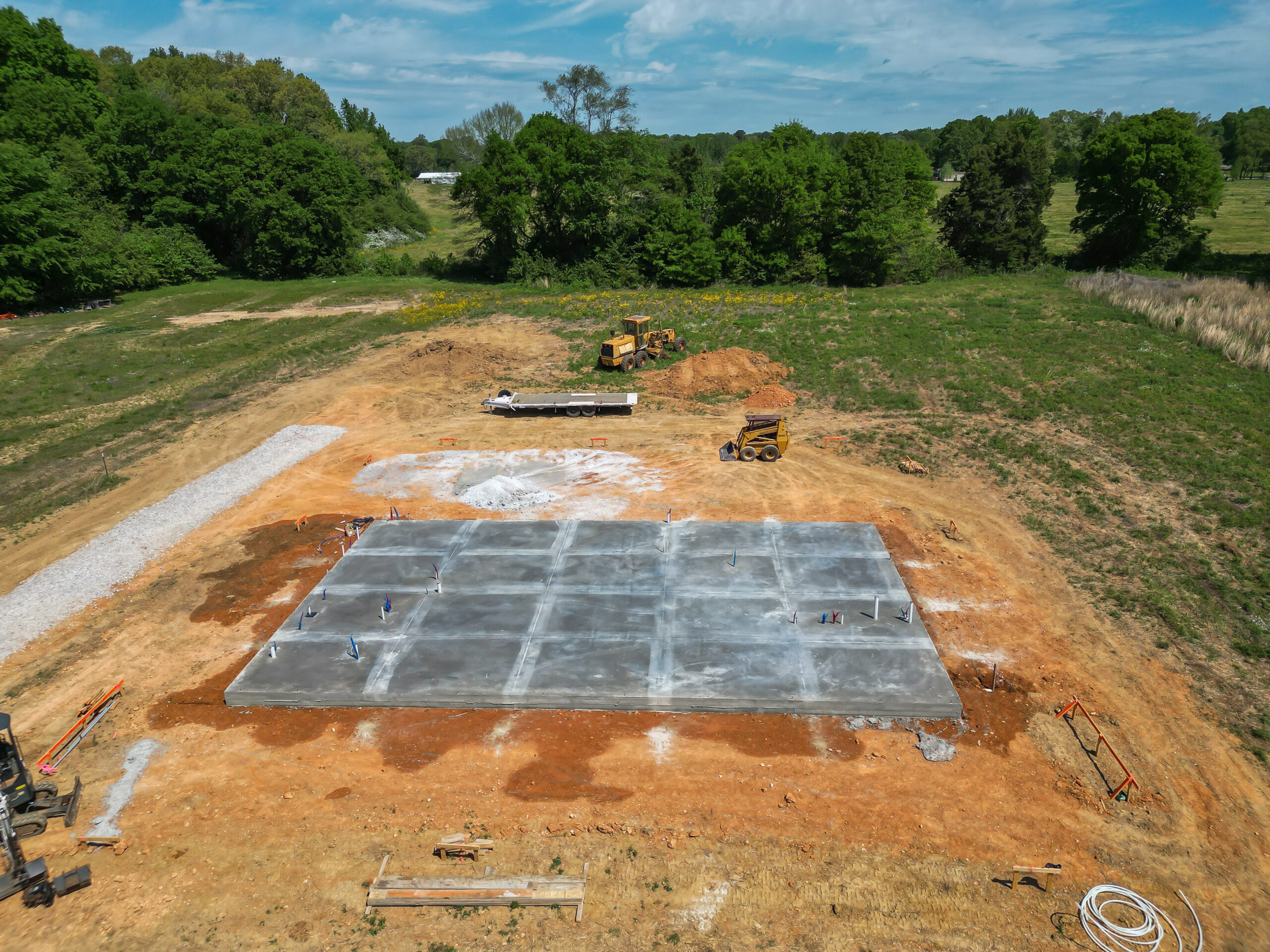 Aerial view of a construction site with a large concrete slab, dirt piles, and a yellow bulldozer near the trees in the background.